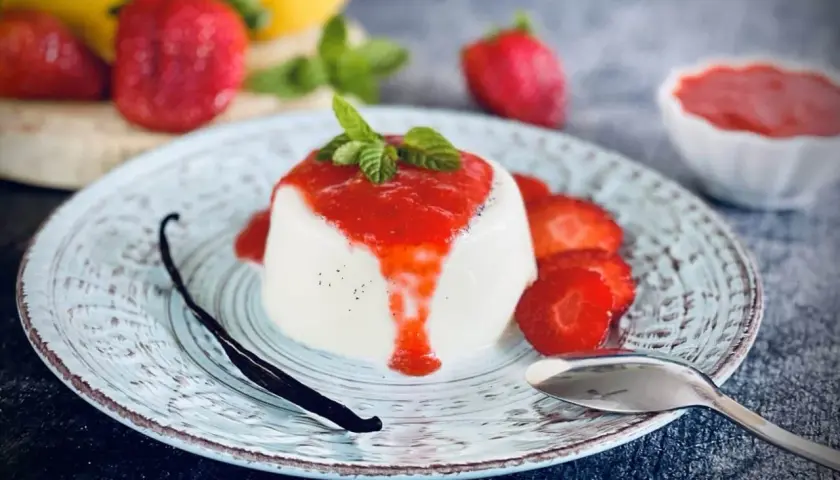 Cremige Panna Cotta mit Erdbeersauce, frischen Erdbeeren, Minze und einer Vanilleschote auf einem Teller.