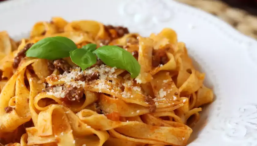 Pyszne tagliatelle bolognese z parmezanem i listkami bazylii na białym talerzu.