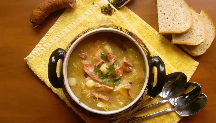 Kapuśniak jak u babci, z kiełbasą i ziemniakami, podany w ozdobnym garnku z chlebem i pieprzem.