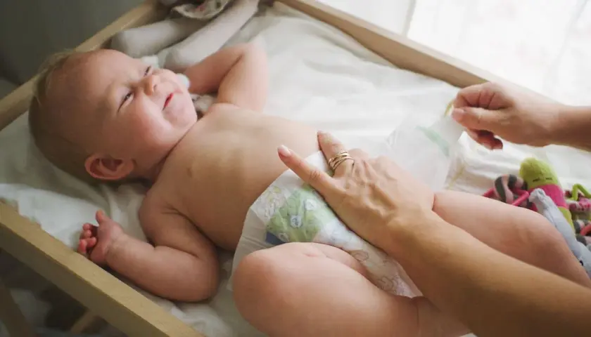 Radosna pielęgnacja niemowlaka chłopca. Mama zmienia pieluszkę, maluch uśmiecha się, leżąc na przewijaku wśród zabawek.