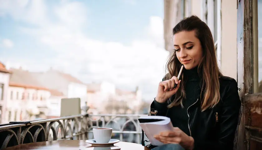 Młoda kobieta z notatnikiem i kawą zastanawia się, czy warto publikować najpierw na platformach typu Wattpad czy Royal Road.