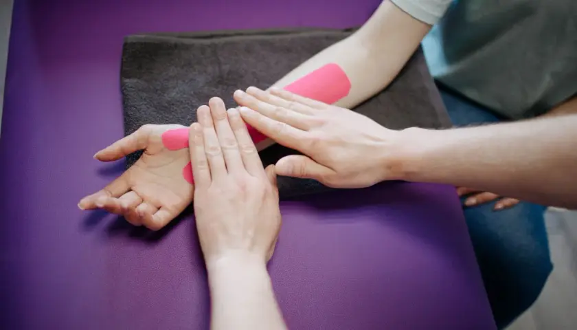 Fizjoterapeuta wykonuje slump test, aplikując różową taśmę kinezjotaping na przedramieniu i dłoni pacjenta.