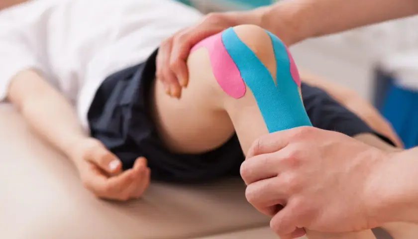 Fizjoterapeuta pokazuje, jak przykleić tejpy na kolano dziecka. Kolorowe plastry zapewniają wsparcie.