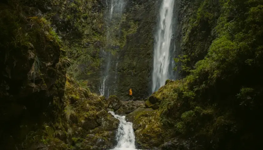 A lone hiker in an orange jacket stands at the base of the majestic Caldeirão Verde waterfall, a breathtaking sight on this lush hike.