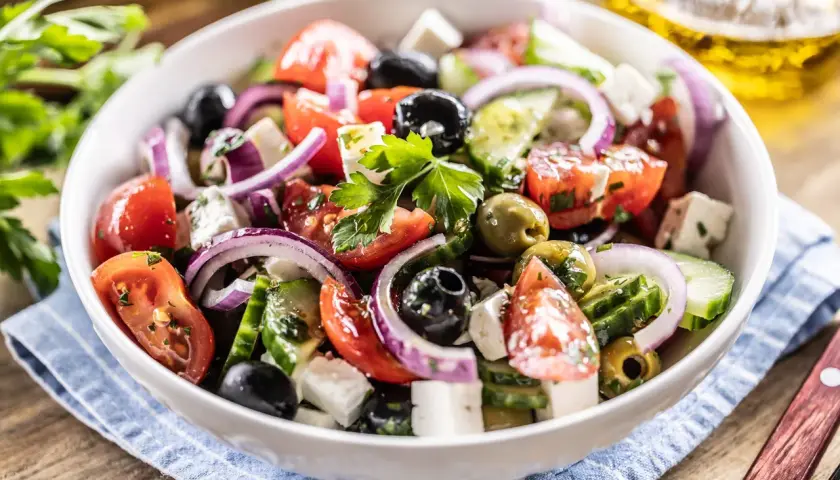 Ensalada griega fresca con tomates, pepinos, cebolla morada, aceitunas negras y queso feta, servida en un bol blanco.