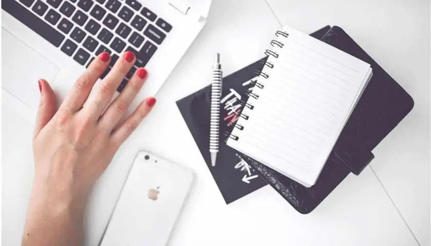 Karta pracy na biurku: dłoń na klawiaturze laptopa, obok telefon, notatnik i długopis.