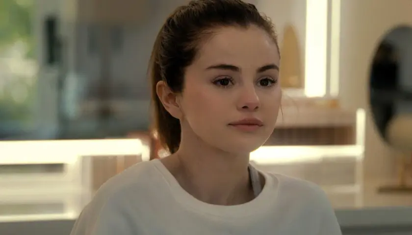 Selena Gomez w białej bluzie, z włosami związanymi w kucyk, patrzy w bok zamyślona.