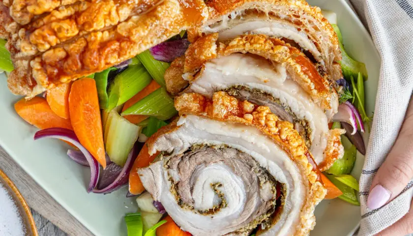 Knusprige Porchetta, ein italienischer Schweinebraten, wird mit karamellisierten Karotten, Sellerie und roten Zwiebeln serviert.