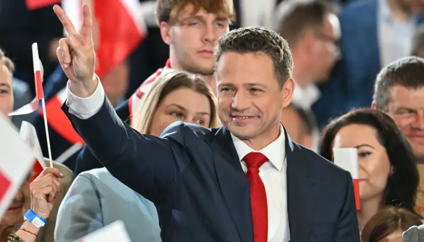Rafał Trzaskowski w garniturze i czerwonym krawacie, pozdrawia tłum, unosząc rękę ze znakiem zwycięstwa.
