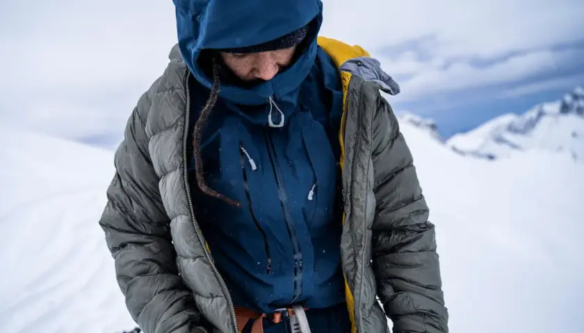 Person in Daunenjacke im Schnee. Die Jacke ist grau, darunter eine blaue Schicht. Perfekt für das Daunenjacke waschen nach einer Bergtour.