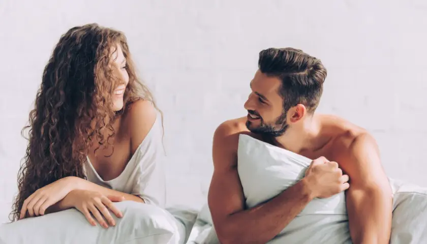Coppia sorridente a letto, con la donna dai capelli ricci che guarda l'uomo con barba, un momento intimo prima di fare sesso.