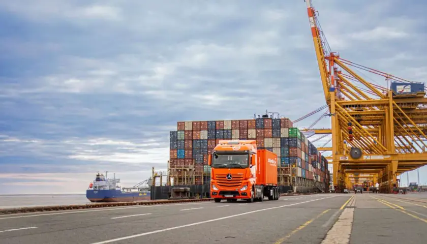 Ein orangefarbener LKW fährt an einem Containerschiff und einem Hafenkranausleger vorbei. Das zeigt, was eine Spedition leistet: Güter transportieren.