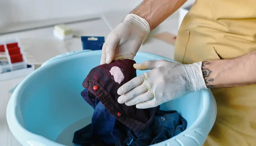 Hände mit Handschuhen behandeln ein Kleidungsstück mit einem rosa Fleck in einer blauen Schüssel, um **Flecken entfernen** zu können.