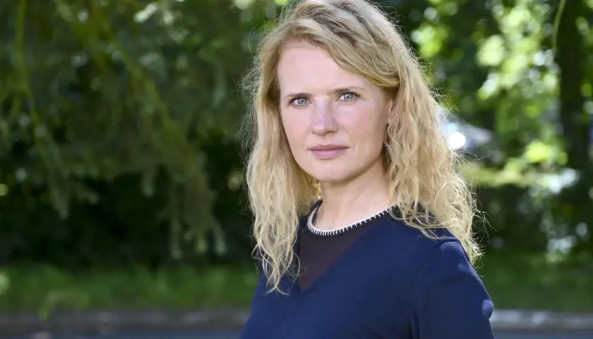 Wiktoria Grodecka z falowanymi blond włosami i niebieskimi oczami, w granatowym swetrze, na tle zieleni.