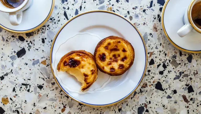 Dwa pyszne pastel de nata na talerzyku, obok filiżanki kawy. Idealny deser na popołudnie.