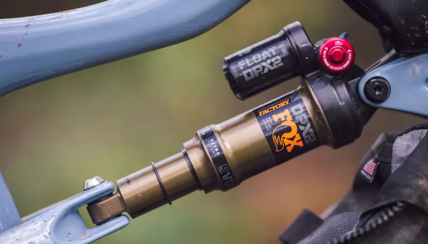 Close-up of a blue bicycle frame with a gold and black Fox Float DPX2 Factory shock. The shock has "FACTORY" and "DPX2" branding.