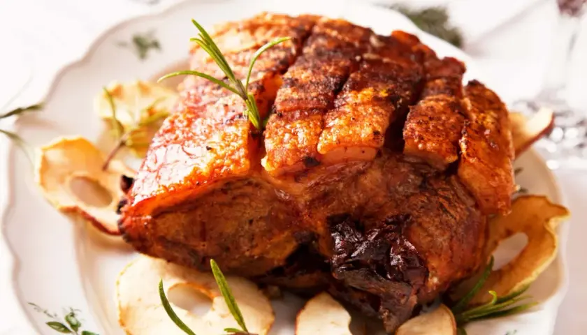 Ein knuspriger Krustenbraten aus dem Backofen, garniert mit Rosmarin und Apfelringen auf einem festlichen Teller.
