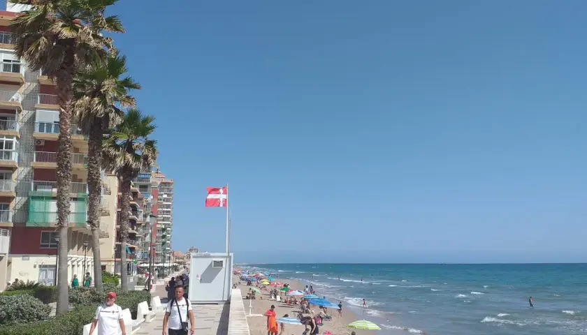Gente disfrutando del sol y el mar en la playa Mareny de Barraquetes, con palmeras y edificios de apartamentos.
