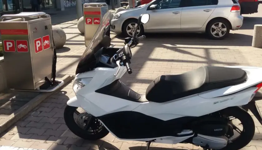 Na parkingu stoi biały skuter. Obok znajduje się automat z oznaczeniami "P" i symbolem motocykla, co sugeruje, że motocykle płacą za parking.
