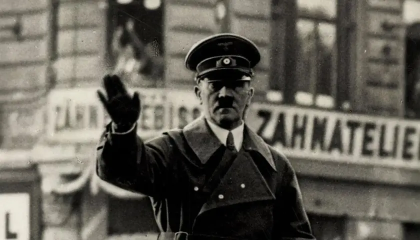 Adolf Hitler winkt. Er trug eine Uniform und einen Hut. Hinter ihm ist eine Tafel mit der Aufschrift "Zahnatelie". Was hat Hitler gemacht? Er hat den Zweiten Weltkrieg begonnen.