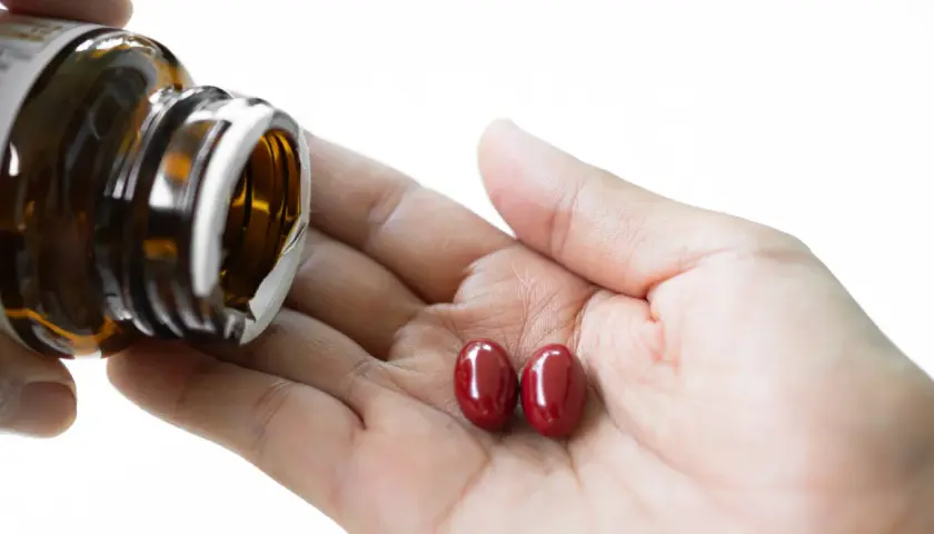 Zwei rote Kapseln liegen in einer Hand, während eine braune Flasche entleert wird. Fragen zur Sicherheit von Astaxanthin krebserregend bleiben offen.