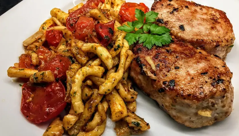 Deux côtelettes de veau grillées, servies avec des pâtes torsadées et des tomates cerises poêlées, garnies de persil frais. Un plat savoureux et coloré.