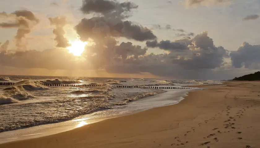 Piękny zachód słońca we Władysławowie. Słońce przebija się przez chmury, tworząc złote refleksy na falach Bałtyku. Na piaszczystej plaży widać ślady stóp.