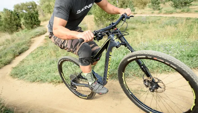 A rider on a black and yellow single fork mountain bike navigates a dusty trail, wearing knee pads and camouflage shorts.