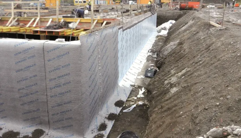 Baustelle mit frisch verlegter Noppenbahn wie rum. Betonfundament und Baugrube sind sichtbar.