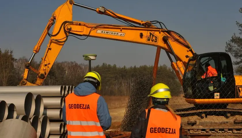 Geodeci w kamizelkach obserwują pracę koparki. Zastanawiasz się, ile kosztuje wynajem koparki?