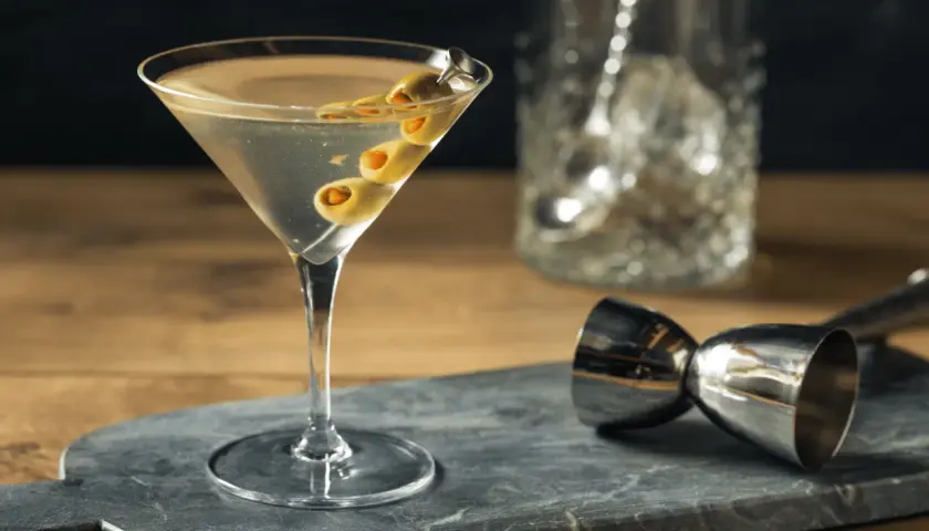 Un verre de martini gin, garni de trois olives, prêt à être dégusté. Un doseur à cocktail et un pichet à glaçons sont visibles en arrière-plan.