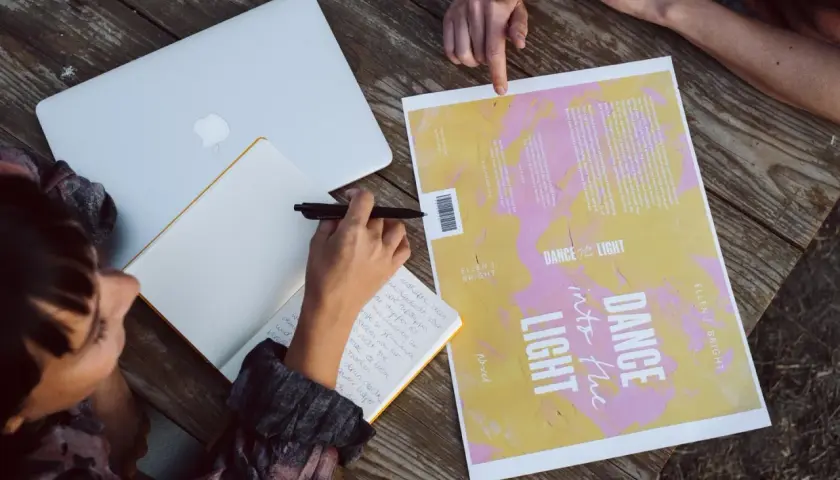 Des mains travaillent à créer une couverture de livre. Un ordinateur portable, un carnet et un brouillon de couverture sont sur une table en bois.