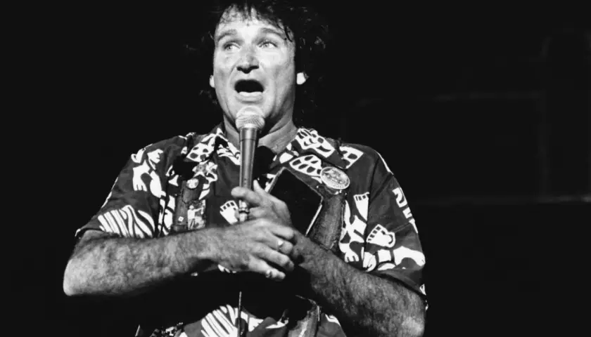 Robin Williams, w hawajskiej koszuli, śpiewa do mikrofonu, trzymając w dłoni notes.