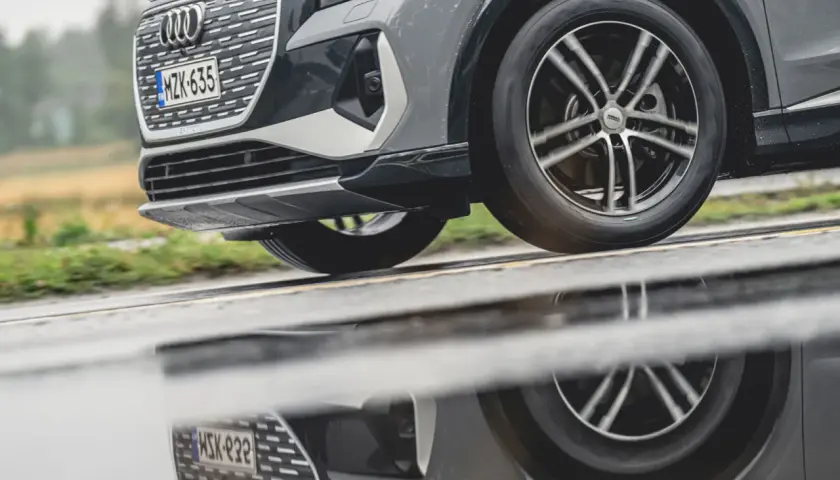 En Audi kör på en regnig väg, dess hjulbultar syns tydligt i den blöta asfalten som reflekterar bilen.