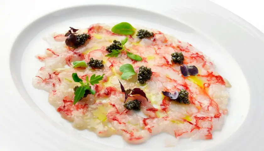 Delicado carpaccio de gambas con caviar, aceite de oliva y brotes verdes, presentado en un plato blanco.