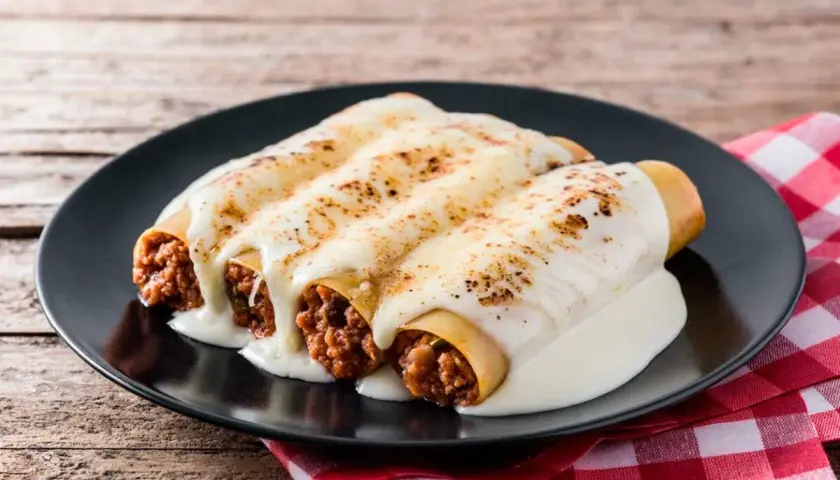 Tres canelones de carne gratinados con queso cremoso, servidos en un plato negro sobre un mantel a cuadros rojos.