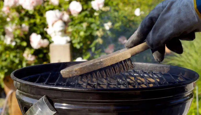 Main tenant gantée, brosse métallique pour nettoyer un barbecue. Des roses roses en arrière-plan.