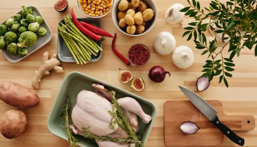 A whole raw chicken in a green dish surrounded by fresh vegetables and herbs on a wooden table.