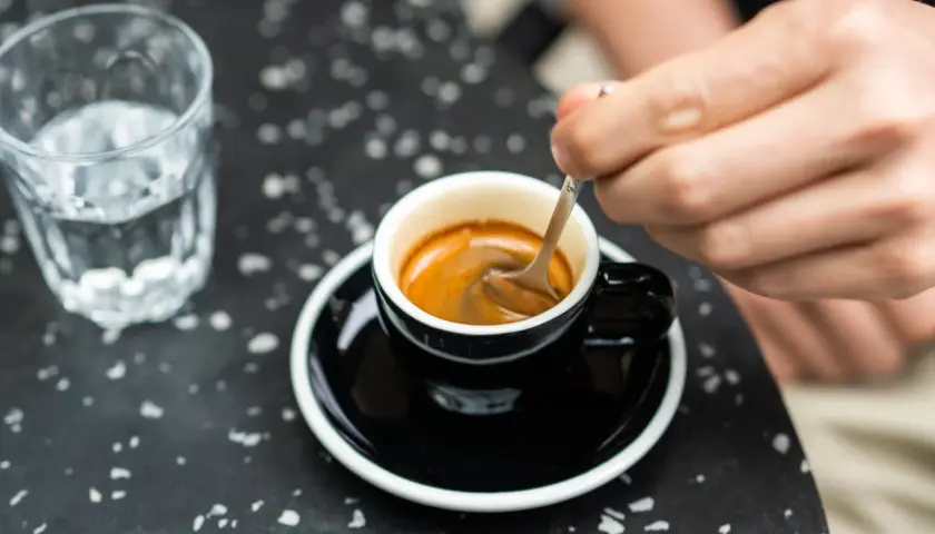 Ręka miesza łyżeczką aromatyczne espresso w czarnej filiżance. Obok szklanka wody. Prosty przepis na espresso jak zrobić.