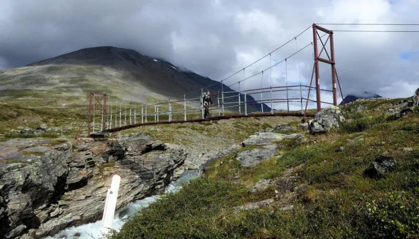 Hängebrucke über einem Fluss im schwedischen Gebirge. Ein Wanderer überquert die Brücke, umgeben von felsiger Landschaft und grüner Vegetation.