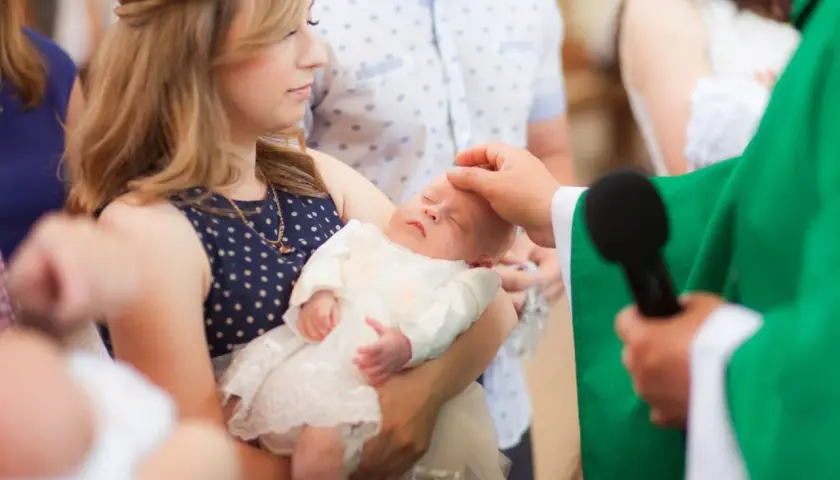 Chrzest dziecka, młoda kobieta trzyma niemowlę, a ksiądz namaszcza je. Czy rodzeństwo może być chrzestnymi?