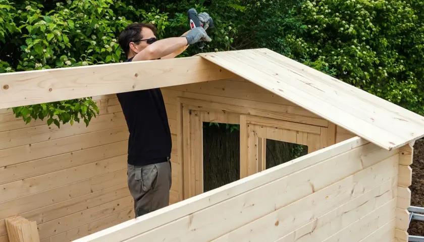 Un homme assemble une cabane en bois, utilisant une perceuse pour fixer une planche sur le toit. La peinture sur bois est visible sur les murs et le toit.