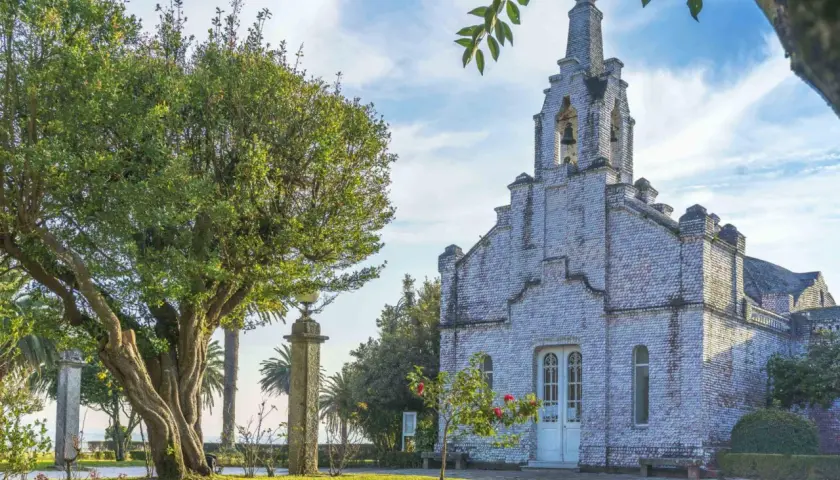 La iglesia de la Toja, cubierta de conchas, se alza majestuosa bajo un cielo azul con nubes. Un árbol frondoso y palmeras la rodean.