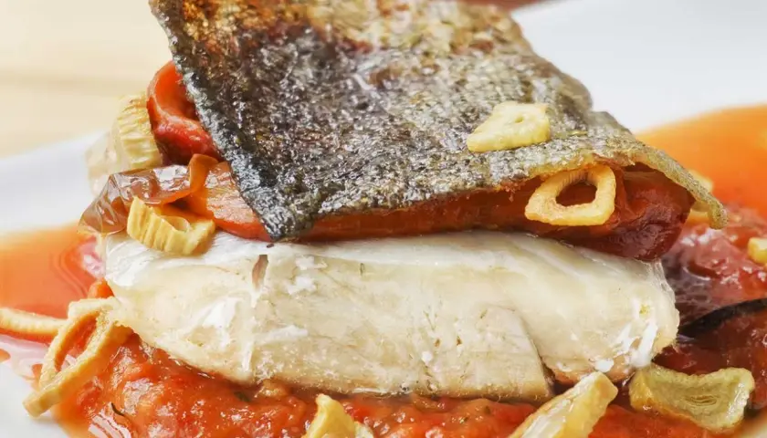 Bacalao a la riojana con piel crujiente, pimientos rojos y ajo frito, servido sobre una cama de salsa de tomate.