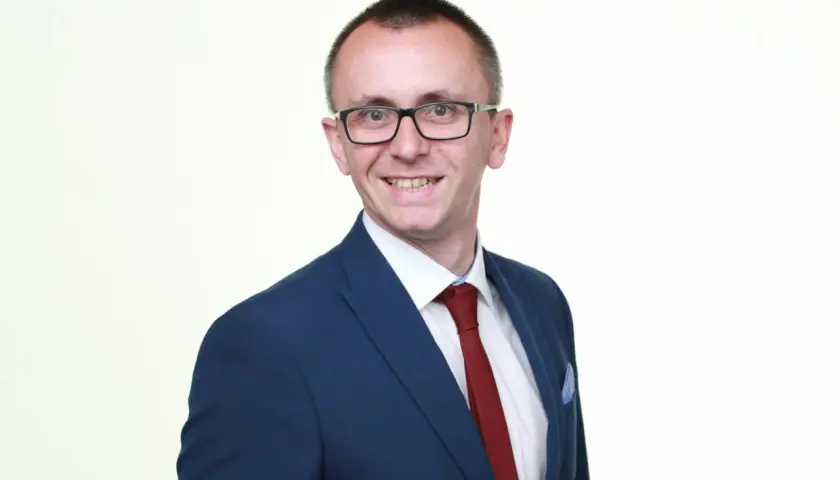 Mężczyzna w okularach i granatowej marynarce z czerwonym krawatem, uśmiecha się.