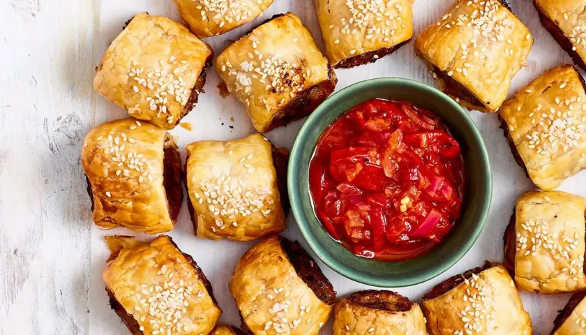 Goldbraune Würstchen im Blätterteig mit Sesam, daneben eine Schale mit roter Chutney-Sauce. Perfekt für schnelle Partysnacks zum Vorbereiten!