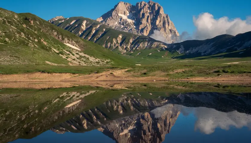 Spiegelglatte See reflektiert majestätisches **Gebirge in Italien** unter blauem Himmel. Grüne Hänge und felsige Gipfel.