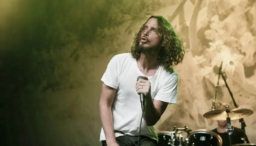 Chris Cornell z zespołem na scenie, w białej koszulce, śpiewa do mikrofonu. W tle perkusja i światła.