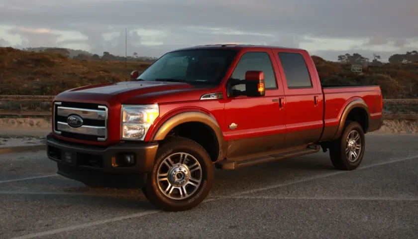 Czerwony Ford F350 King Ranch z brązowym pasem, zaparkowany na tle krajobrazu z wydmami i drewnianym płotem.