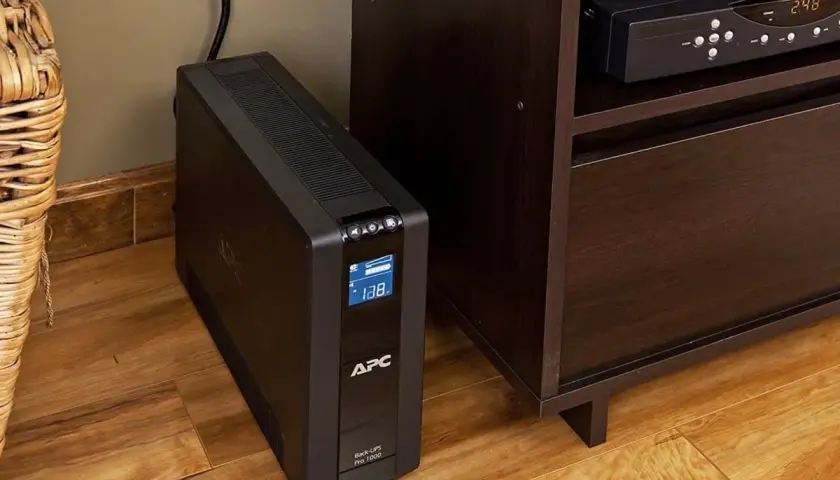 Ups co to? Czarny UPS APC Back-UPS Pro 1000 stoi na drewnianej podłodze obok mebla.
